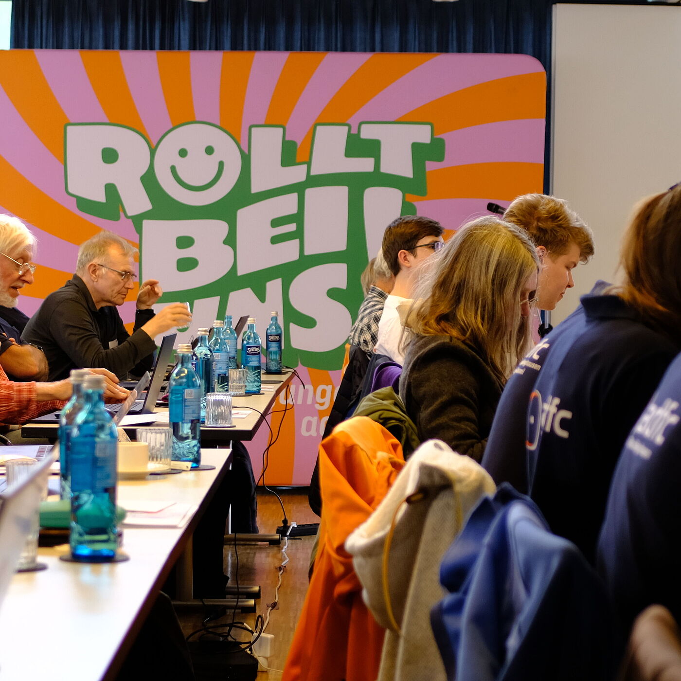 2026_04_18_Landesversammlung_Muenster_ADFC-NRW Mehrere Personen, Stuhlreihen mit Plakat im Hintergrund.