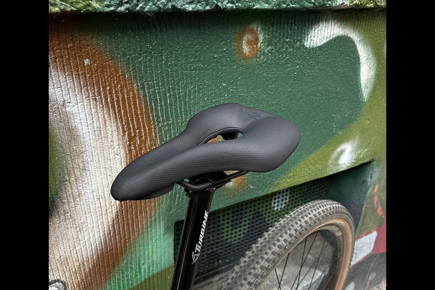 Fingerprint Seite Schwarzer Fahrradsattel mit mittlerer Aussparung, montiert an einem Fahrrad vor einer bemalten Wand.